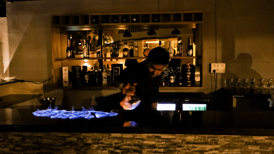 A man behind a counter table with drinks on the shelf behind him at Amara Grand Baga, Goa.